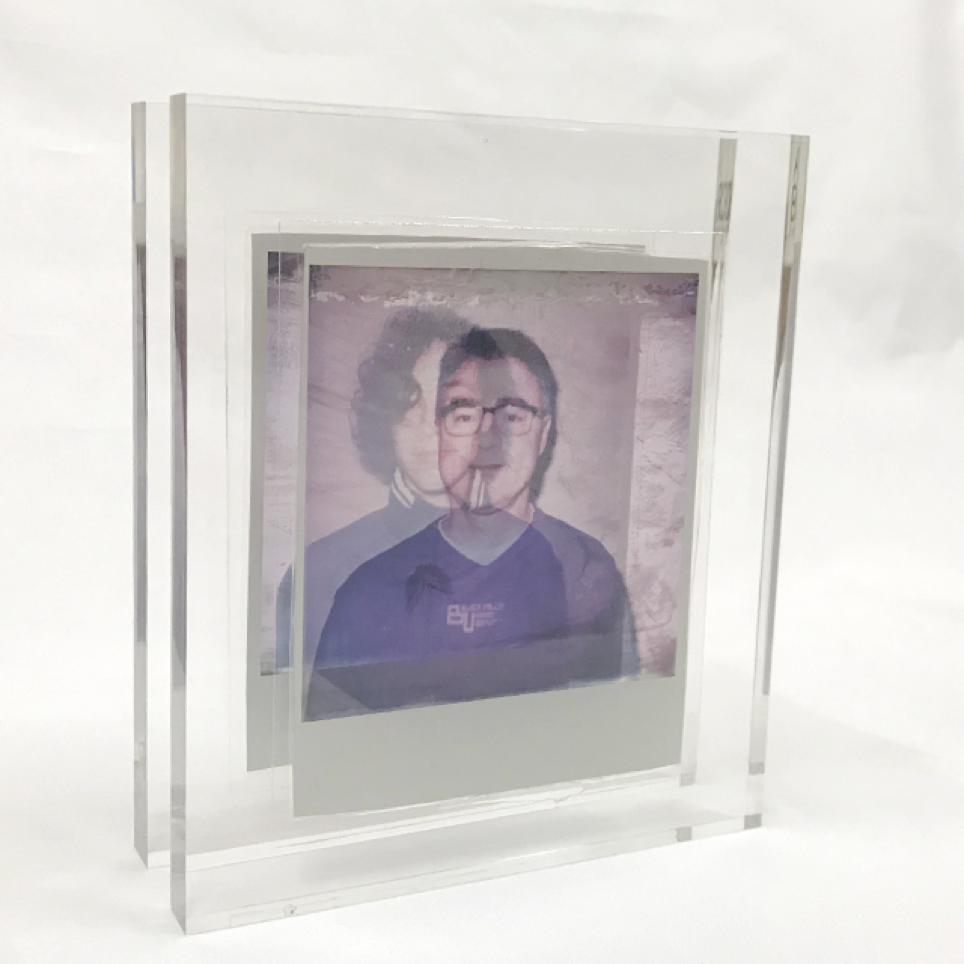 A picture of polaroid peels encased in two separate transparent acrylics. Because the polaroids are peeled, they are also transparent. 
The two acrylic cases are placed in font of each other. In the front the portrait of a father is shown. Behind the portrait of his son. The acrylics are set up in a diagonal line, so it shows the space between the two.