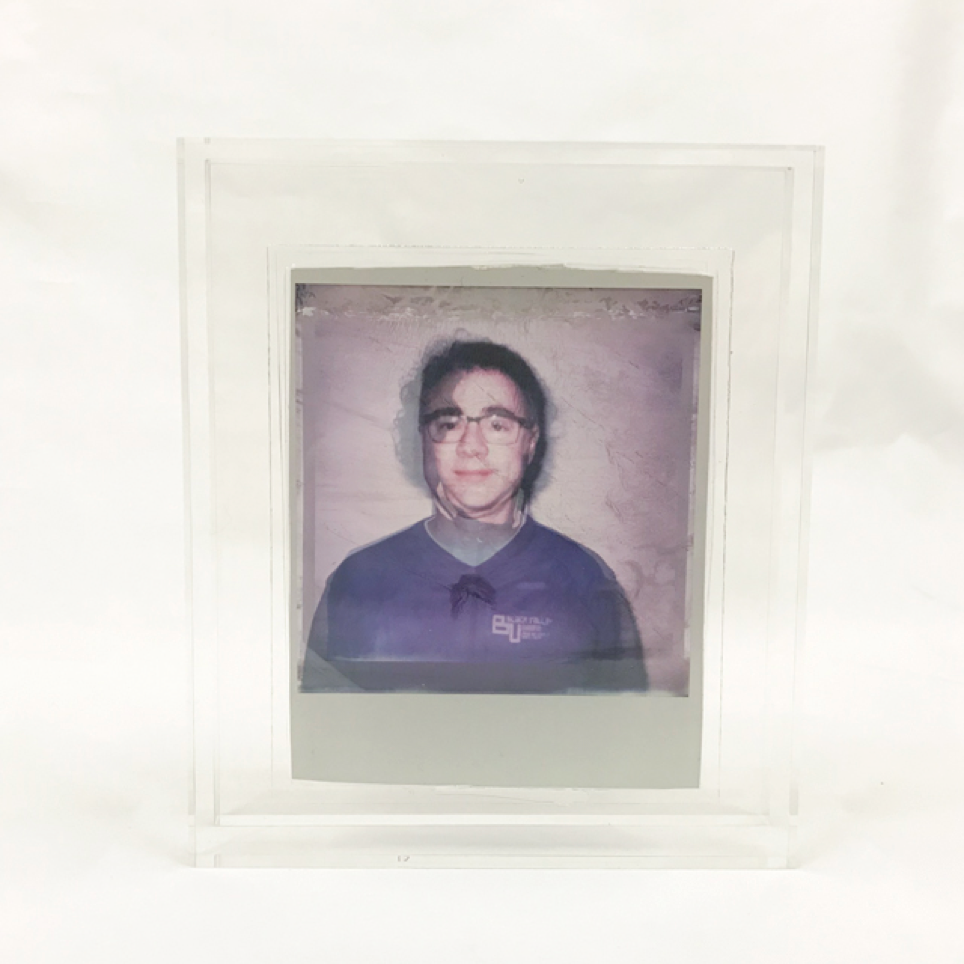 A picture of polaroid peels encased in two separate transparent acrylics. Because the polaroids are peeled, they are also transparent. 
The two acrylic cases are placed in font of each other. In the front the portrait of a father is shown. Behind the portrait of his son. The acrylics are set up in such a way the two merge into one, showing one face. The emphasises the features they share. 