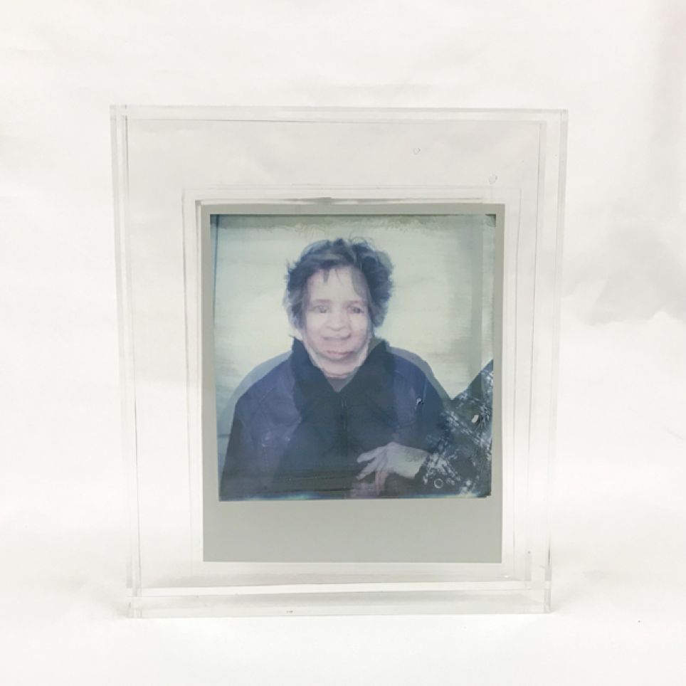 A picture of polaroid peels encased in two separate transparent acrylics. Because the polaroids are peeled, they are also transparent. 
The two acrylic cases are placed in font of each other. In the front the portrait of a grandmother is shown. Behind the portrait of her granddaughter. The acrylics are set up in such a way the two merge into one, showing one face. The emphasises the features they share. 